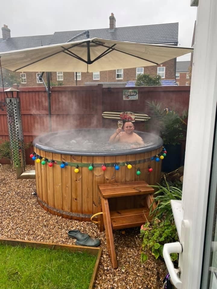 Gardenvity wood fired hot tub with steam rising in small backyard surrounded by wooden fence.