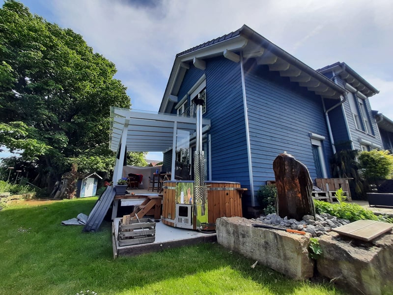 Gardenvity wood fired hot tub next to blue wooden house in rural landscape.
