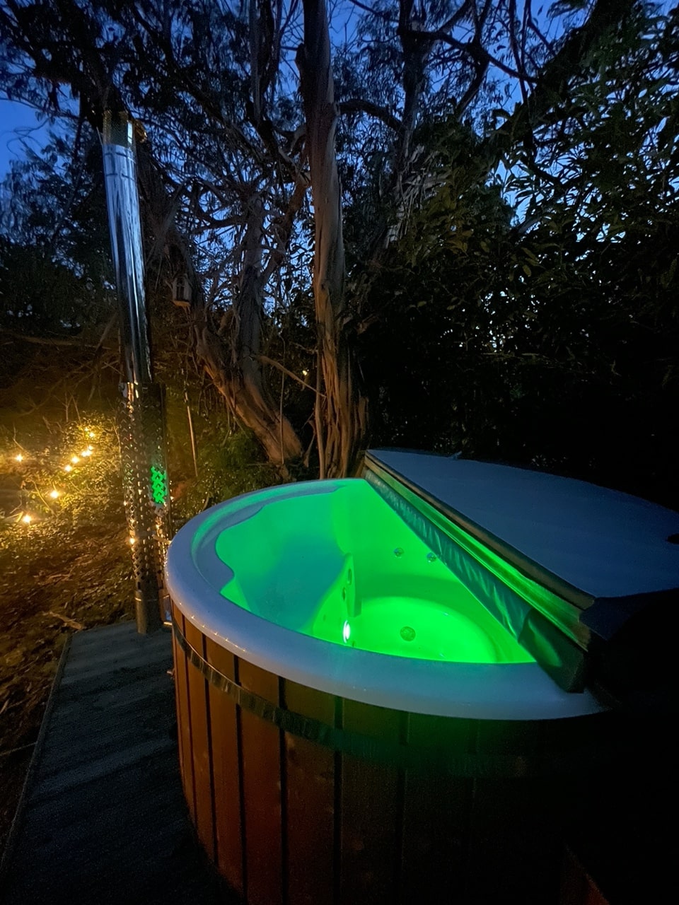 Green-lit Gardenvity wood fired hot tub in secluded garden surrounded by trees.